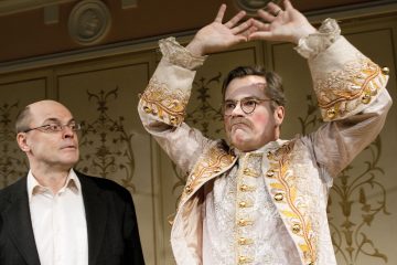 Lavalla on kaksi miestä; toinen mustaan pukuun pukeutuneista näyttää yllättyneeltä, kun taas toinen, jolla on koristeellinen, kirjailtu puku, jossa on kultainen koristelu, nostaa dramaattisesti kätensä ylös, ja hänellä on vakava ilme ja maalatut viikset.