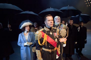 Sotilaspukuinen mies seisoo juhlallisesti BBC:n vanhanajan mikrofonin ääressä, ja hänen ympärillään on juhlapukuisia ihmisiä, joilla on kädessään mustat sateenvarjot. Siniseen takkiin ja leveälieriseen hattuun pukeutunut nainen seisoo lähellä ja näyttää huolestuneelta.