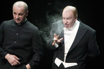 Kaksi tummiin pukuihin pukeutunutta miestä seisoo vierekkäin; toisella on papin kauluspaita, kun taas toisella on valkoinen rusetti ja smokki, ja hänellä on sytytetty sikari, jonka savu kiertää hänen ympärillään mustaa taustaa vasten.