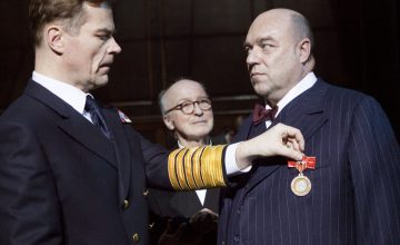 Tummiin univormuihin pukeutunut mies kiinnittää mitalin toiselle miehelle, jolla on nastaraitainen puku ja rusetti, ja kolmas silmälasipäinen mies katselee taustalla.