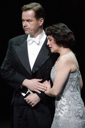 Mustaan smokkiin pukeutunut mies ja hopeanhohtoiseen mekkoon pukeutunut nainen seisovat lavalla lähekkäin ja näyttävät synkiltä ja tunteikkailta tummaa taustaa vasten.