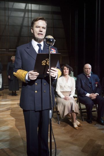 Merivoimien univormuun pukeutunut mies seisoo vintage-mikrofonin ääressä ja lukee kansiosta, kun hänen takanaan lavalla istuu kolme viralliseen asuun pukeutunutta henkilöä hämärässä teatteriympäristössä.