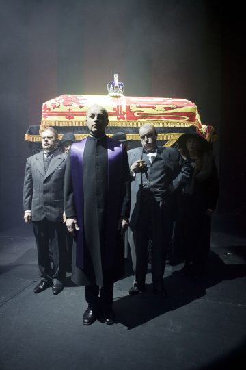 Neljä viralliseen asuun pukeutunutta ihmistä kantaa juhlallisesti kuninkaallista, lippuun verhottua arkkua, jonka päällä on kruunu, dramaattisessa valaistuksessa ja hämärässä taustalla, joka tuo mieleen synkän hautajaiskohtauksen.