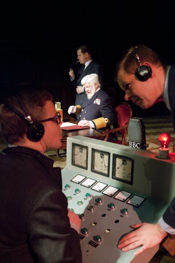 Neljä vintage-vaatteisiin pukeutunutta miestä on mukana radiolähetysasetelmassa. Yksi käyttää ohjauspaneelia, toinen kumartuu puhumaan, kolmas istuu pöydän ääressä kuulokkeet päässä ja neljäs seisoo takana ja puhuu mikrofoniin.