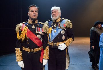 Lavalla seisoo vierekkäin kaksi miestä koristeellisissa sotilasunivormuissa, joissa on mitalit, vyötäröt ja kultaiset olkaimet. Molemmat näyttävät vakavilta; toisella on punainen ja toisella sininen liivi. Taustalla on tummiin vaatteisiin pukeutuneita ihmisiä.