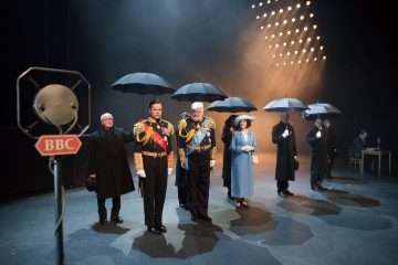 Aikakauden pukuihin pukeutuneet näyttelijät seisovat lavalla rivissä mustat sateenvarjot kädessään, etualalla on vanha BBC:n mikrofoni ja dramaattinen näyttämövalaistus valaisee kohtauksen.