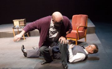 Kalju mies, jolla on violetti neuletakki, polvistuu lattialla makaavan pukumiehen päälle ja näyttää pidättelevän häntä lavalla, jonka taustalla on punainen nojatuoli ja pieni pöytä.