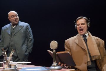 Kaksi miestä lavalla: toinen seisoo puvussaan ja katselee, kun taas toinen istuu pöydän ääressä mikrofonin, kuulokkeiden ja kansion kanssa ja näyttää puhuvan tai laulavan intohimoisesti. Ympäristö on hämärästi valaistu ja teatraalinen.