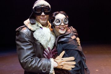 Kaksi ihmistä vintage-lentäjien asuissa ja suojalaseissa hymyilee ja poseeraa yhdessä pimeällä näyttämöllä, toinen nahkatakki ja toinen viitta päällään, mikä viittaa leikkisään tai teatterikohtaukseen.