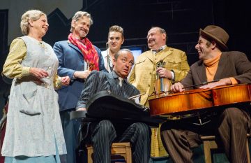 Kuusi värikkäisiin, vintage-tyylisiin pukuihin pukeutunutta ihmistä seisoo yhdessä hymyillen ja nauraen. Kaksi miestä istuu soittimiensa kanssa, kun muut seisovat heidän ympärillään ja näyttävät nauttivan vilkkaasta hetkestä lavalla.