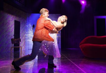 Punapaitainen mies kastaa valkoiseen mekkoon pukeutuneen naisen tanssiessaan kirkkaasti valaistulla lavalla, jonka taustalla on violetti valaistus, punainen sohva ja tuoli.