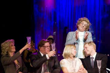 Pöydän ääressä istuu viisi juhlapukuista ihmistä, joista neljä nostaa lasit maljan kohotukseksi, kun yksi nainen seisoo heidän takanaan taputtaen ja hymyillen eloisan sinistä näyttämötaustaa vasten.