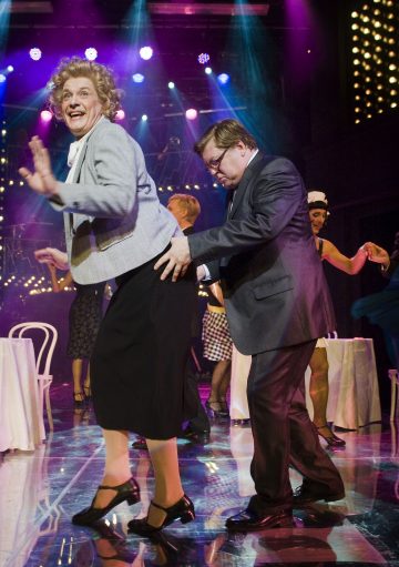 Pukumies tanssii leikkisästi vanhemmaksi naiseksi pukeutuneen henkilön takana kirkkaasti valaistulla näyttämöllä, jonka taustalla on muita esiintyjiä ja valkoisia tuoleja.