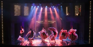 Kuusi värikkäisiin pukuihin pukeutunutta tanssijaa esittää korkealta potkivan can-can-rutiinin kirkkaasti valaistulla näyttämöllä, jota ympäröivät eloisat valonheittimet ja valaistut telttavalot.