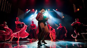 Lavalla laulaa punaisessa takissa ja verkkosukissa esiintyvä drag-esiintyjä, jota ympäröivät can-can-tyylisiin pukuihin pukeutuneet tanssijat dramaattisessa sinipunaisessa valaistuksessa.