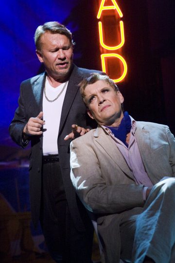 Kaksi miestä lavalla, toinen seisoo ja puhuu sikari kädessään, puku ja valkoinen t-paita, toinen istuu vaaleassa puvussa ja näyttää mietteliäältä. Heidän takanaan loistaa neonkyltti, jossa lukee AUD.