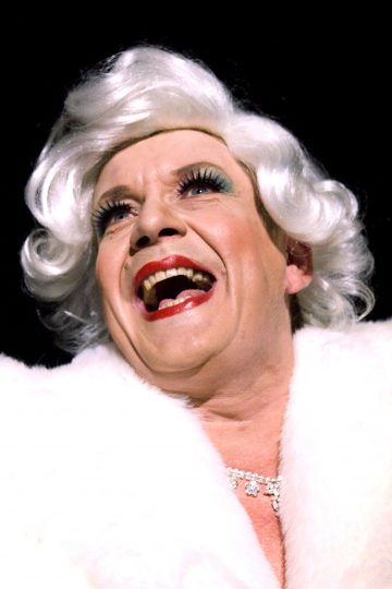 Henkilö, jolla on dramaattinen meikki, kirkkaanpunainen huulipuna ja tekoripset, hymyilee leveästi, kun hänellä on valkoinen peruukki, valkoinen turkistakki ja kimalteleva kaulakoru mustaa taustaa vasten.