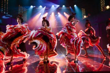 Tanssijat tanssivat can-cania lavalla värikkäiden näyttämövalojen alla punaisissa, valkoisissa ja vaaleanpunaisissa puvuissa, joissa on röyhelöhameet. Kohtaus on vilkas ja energinen.