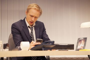 Pukumies istuu työpöydän ääressä ja katsoo yllättyneenä puhelintaan. Pöydällä on toimistopuhelin, kuittitulostin, lamppu ja kehystetty valokuva lapsesta. Pystysuorat kaihtimet peittävät taustan.