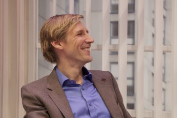 Vaaleatukkainen mies, jolla on harmaa bleiseri ja sininen paita, hymyilee ja katsoo vasemmalle istuen ikkunan edessä, jossa on pystysuorat kaihtimet.