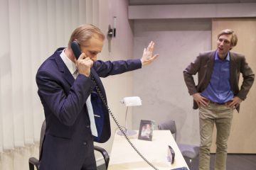 Pukumies puhuu pöydän ääressä olevaan langalliseen puhelimeen ja osoittaa kädellään pysähtymistä toiselle lähellä seisovalle miehelle, joka vaikuttaa turhautuneelta. Työpöydällä on lamppu, valokuvakehys ja toimistotarvikkeita.