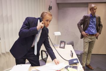 Pukumies puhuu johtoon kytkettyyn puhelimeen ja katsoo samalla pöydällä olevia papereita modernissa toimistossa. Toinen bleiseriasuinen mies seisoo lähellä ja tarkkailee häntä kädet taskuissaan.