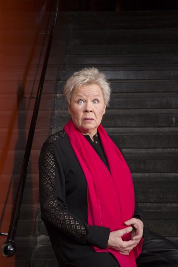 Vanhempi nainen, jolla on lyhyet harmaat hiukset, on pukeutunut mustaan asuun ja kirkkaan vaaleanpunaiseen huiviin, seisoo tummia portaita vasten, toinen käsi vatsallaan, ja katsoo suoraan kameraan vakavalla ilmeellä.