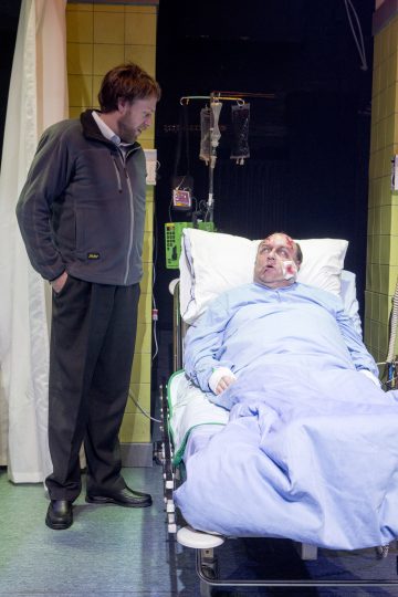 Tummiin takkeihin pukeutunut mies seisoo sairaalasängyn vieressä ja katselee toista miestä, joka makaa sängyssä siteet kasvoillaan ja lääkintäletkut kiinnitettyinä, kliinisessä ympäristössä.