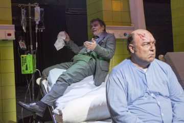 Kaksi miestä sairaalahuoneessa; toinen pukumies istuu sairaalasängyllä ja viittoo nenäliinalla, kun taas toinen istuu etualalla sängyllä siniseen pyjamaan pukeutuneena ja päävammaisena, vakavana. Lääkinnälliset laitteet ovat näkyvissä.