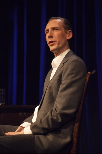 Harmaaseen pukuun ja valkoiseen paitaan pukeutunut mies istuu puisella tuolilla, puhuu tai esiintyy lavalla, jonka taustalla on siniset verhot.