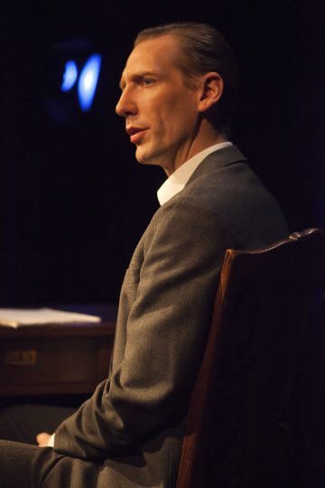 Pukumies istuu puisen työpöydän ääressä sivuttain ja puhuu tai esittää hämärässä ympäristössä, mahdollisesti näyttämöllä, jonka taustalla on teatterivalaistus.