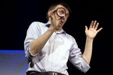 Vaaleansiniseen paitaan pukeutunut henkilö pitää teippirullaa toista silmäänsä vasten ja tekee yllättyneen ilmeen suurilla silmillä ja kohotetuilla kulmakarvoilla, samalla kun hän nostaa toisen kätensä ilmaan tummaa taustaa vasten.