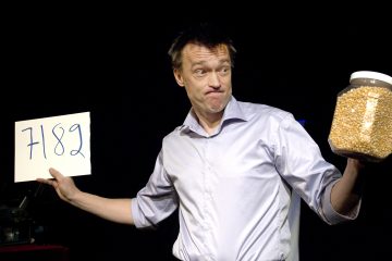 Vaaleaan paitaan pukeutunut mies pitää toisessa kädessään valkoista korttia, johon on kirjoitettu 7182, ja toisessa kädessään suurta purkkia popcorn-ytimiä, ja seisoo tummalla taustalla leikkisästi.