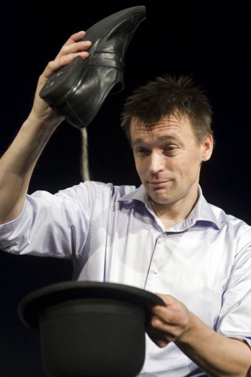 Valkoiseen paitaan pukeutunut mies pitää mustaa kenkää ylösalaisin mustan silinterihatun päällä ja näyttää yllättyneeltä kuin taikatemppua tehneeltä, tummaa taustaa vasten.