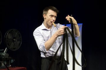 Valkoiseen paitaan pukeutunut mies tutkii sotkeutuneita filmirullia sikari suussaan ja seisoo elokuvaprojektorin vieressä tummaa taustaa vasten.