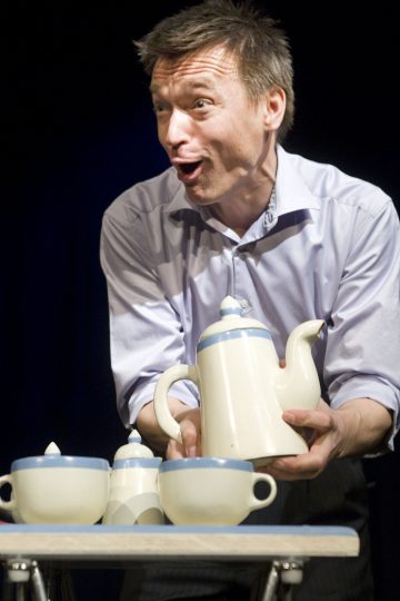 Mies, jolla on eloisa ilme, pitää kädessään valkoista teekannua, jossa on sininen reunus, ja seisoo pöydän takana, jossa on yhteensopivia kuppeja ja sokerikulho, mikä viittaa siihen, että hän esiintyy tai kertoo tarinaa.