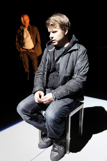 Tummiin takkeihin pukeutunut mies istuu tuolilla valonheittimen alla ja katsoo mietteliäästi alaspäin, kun taas toinen mies seisoo taustalla, myös valaistuna, kädet lanteilla. Muu osa kohtauksesta on pimeä.