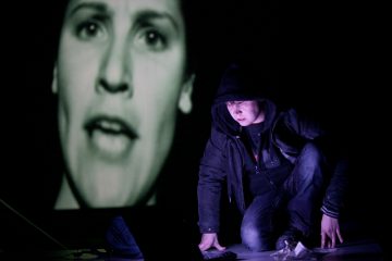 Hupparitakkinen henkilö kyyristelee maassa pimeässä huoneessa, ja hänen takanaan on valaiseva näyttö, jossa näkyy suuri mustavalkoinen lähikuva naisen kasvoista.