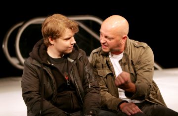 Kaksi miestä istuu tiiviisti yhdessä lavalla. Toinen, jolla on tumma takki päällään, katsoo mietteliäästi toista, joka on kalju ja elehtii kiihkeästi nyrkillä, jolla on vihreä takki päällään. He näyttävät käyvän kiivasta keskustelua.