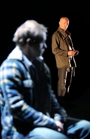 Etualalla istuu ruudulliseen takkiin pukeutunut mies, joka on sumea, kun taas taustalla seisoo vihreään takkiin pukeutunut kalju mies, joka katsoo tarkkaan eteenpäin tummaa taustaa vasten.