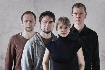 Neljä aikuista seisoo lähekkäin yksinkertaisen, kuvioidun seinän edessä. Kolme miestä ja yksi nainen katsovat kameraan neutraalilla ilmeellä, ja heillä on yllään maanvärisiä ja mustia rentoja vaatteita.