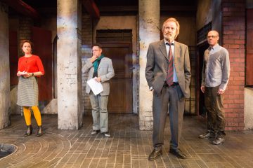 Neljä ihmistä seisoo dramaattisessa poseerauksessa lavalla, jossa on tiili- ja pylväsmainen tausta; kahdella miehellä on puvut, yhdellä miehellä harmaa paita ja tummat housut, ja naisella on punainen yläosa, harmaa hame ja keltaiset sukkahousut.