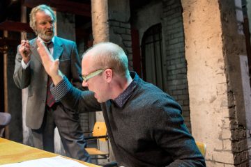 Silmälasipäinen mies istuu pöydän ääressä, nostaa kätensä ylös ja huutaa, kun taas toinen pukumies seisoo hänen takanaan ja katsoo vakavana teollisen näköisessä huoneessa, jossa on tiilipilareita.