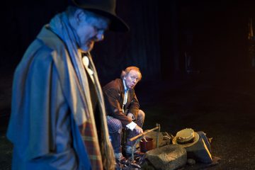 Kaksi näyttelijää esiintyy hämärästi valaistulla näyttämöllä. Toisella etualalla olevalla miehellä on hattu ja takki päässään ja hän katsoo alaspäin. Toinen mies kyyristelee kivien ja rekvisiitan lähellä ja katsoo keskittyneesti kohti ensimmäistä miestä. Kohtaus tuntuu dramaattiselta ja jännittyneeltä.