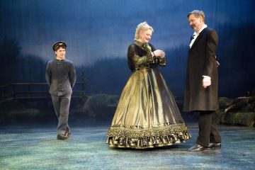 Lavalla esiintyy kolme näyttelijää aikakauden puvuissa; nainen koristeellisessa puvussa seisoo kahden miehen välissä, joista toisella on puku ja toisella univormu, ja heidän takanaan on maalattu silta ja puita.