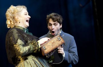 Aikakauden asuun pukeutunut nainen nauraa pitelemällä vanhaa puulaatikkoa, kun harmaaseen takkiin pukeutunut yllättynyt nuori mies katselee sitä suu auki hämmästyksestä.