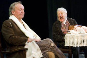 Kaksi iäkästä miestä istuu pitsikankaalla peitetyn pöydän ääressä; toinen näyttää rauhalliselta ja mietteliäältä, kun taas toinen, jolla on ilmeikkäät kasvot, näyttää huutavan tai reagoivan tunteisiin. Kohtaus on asetettu näyttämölle.