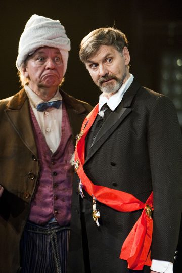 Kaksi teatteriasuihin pukeutunutta miestä seisoo yhdessä; toisella on turbaanimainen päähine, violetti liivi ja rusetti, kun taas toisella on vyö ja muodollinen puku, molemmilla ilmeikkäät kasvot, hämärästi valaistulla näyttämöllä.