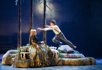 Pitkään mekkoon pukeutunut nainen istuu penkillä metsäisessä lavastuksessa, kun paitaan ja sukkanauhoihin pukeutunut mies hyppää dramaattisesti ilmassa häntä kohti. Kohtaus on teatraalinen ja kirkkaasti valaistu.