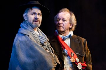 Lavalla seisoo kaksi vanhempaa miestä aikakauden puvuissa; toisella on tumma hattu, huivi ja viitta, kun taas toisella on mitalein koristeltu vyö ja rusetti, ja molemmat vaikuttavat vakavilta ja mietteliäiltä.